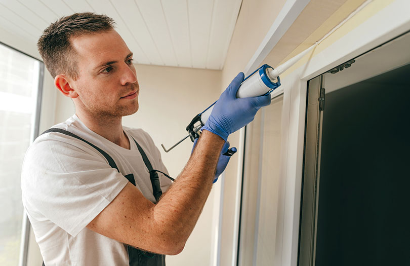 Young man wearing overalls sealing cracks between window and trim using waterproof silicone caulk on the balcony.
